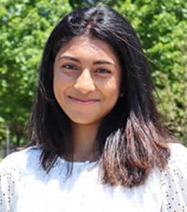 Headshot of an Indian woman smiling. She has shoulder length black hair and is wearing a white long sleeve shirt. Trees are behind her in the background